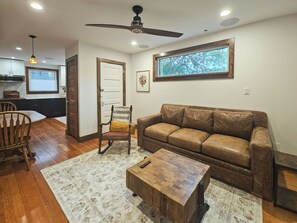 A living room with a brown leather sofa, a wooden coffee table, a wooden chair, a patterned rug, a ceiling fan, and a window with a view of trees.