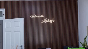 Uma área de recepção com painel de parede de madeira, porta branca, cadeira preta, mesa verde e uma placa com a inscrição 'Welcome to Makay's'.