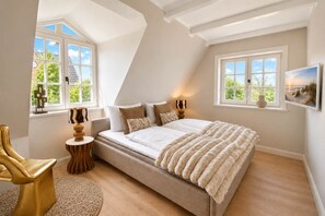 Ein Schlafzimmer mit einem großen Bett, zwei Fenstern, einem Sessel, einer Lampe und einem Bild an der Wand.