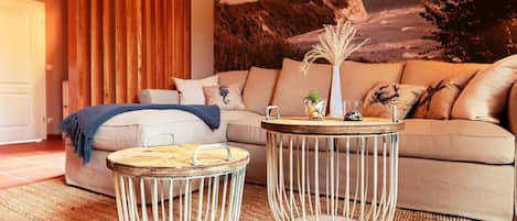 Una sala de estar con un sofá color beige, dos mesas redondas con estructura de metal, un tapiz tejido y un mural de paisaje costero en la pared.