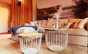Ein Wohnzimmer mit einem beigefarbenen Sofa, zwei runden Tischen mit Metallgestellen, einem Teppich und einem Wandgemälde mit einer Küstenlandschaft.