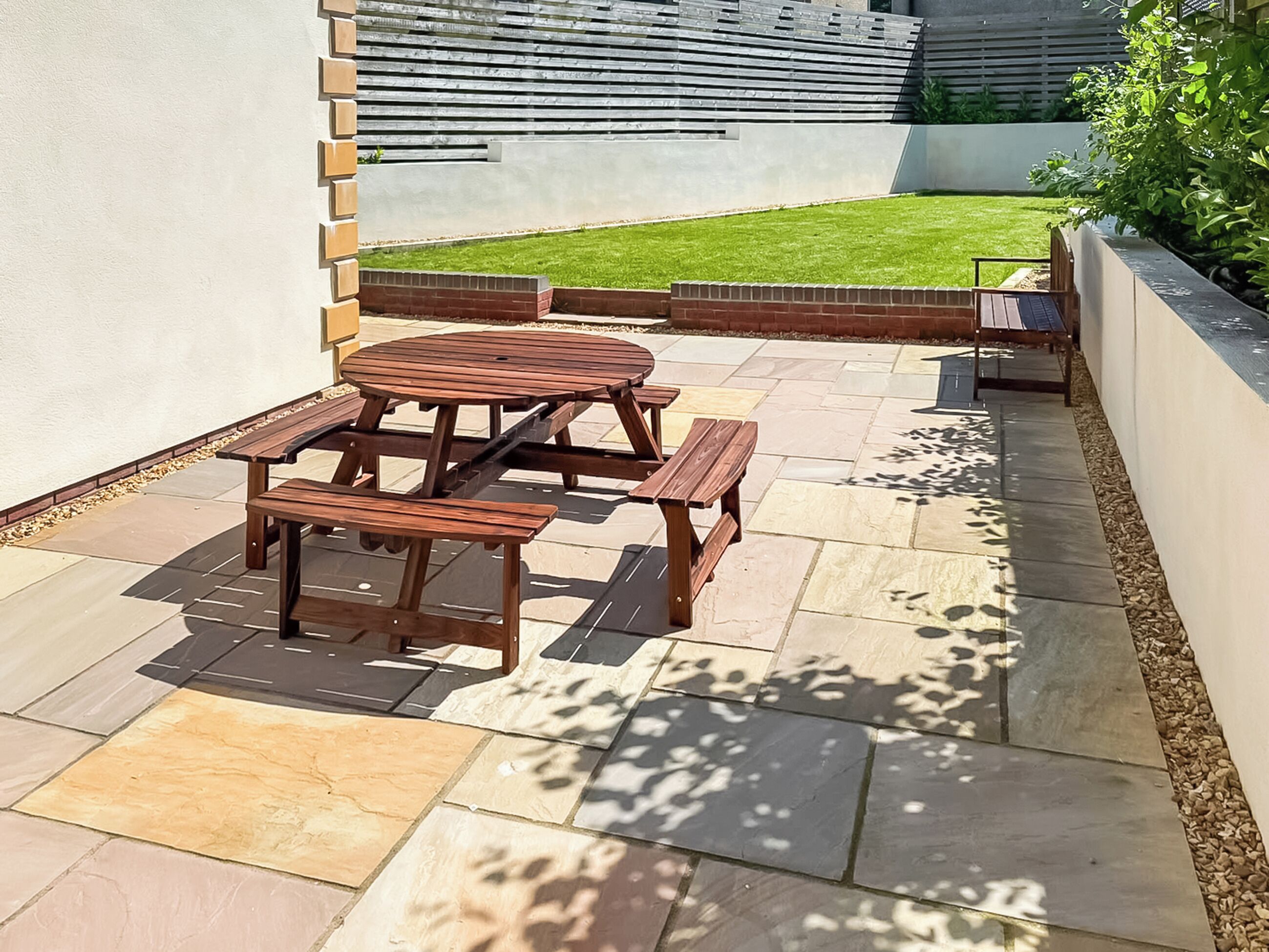 A patio with a wooden table and benches, surrounded by stone tiles and a grassy lawn.