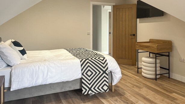 A bedroom with a bed, a wooden door, a TV mounted on the wall, and a small wooden table with a stool. - Barncroft Retreat - UK51693 (High Seaton, near Workington)