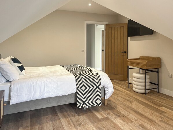 A bedroom with a bed, a wooden door, a TV mounted on the wall, and a small wooden table with a stool.
