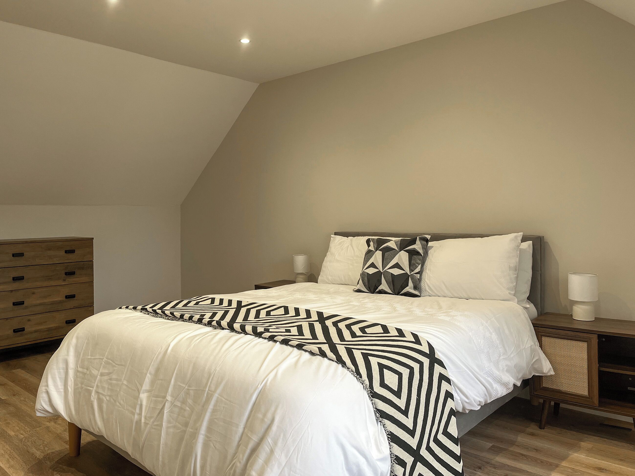 Een slaapkamer met een bed, nachtlampjes, een dressoir en een schuine kap.