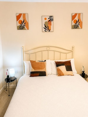 A neatly made bed with a white comforter and brown pillows, a white metal headboard, and three abstract art pieces on the wall.