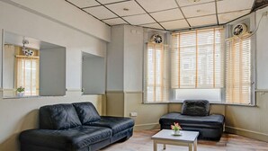 A living room with a black leather sofa, a small table with a flower vase, a mirror, and a window with blinds. - Standard Triple Room (Cardiff)