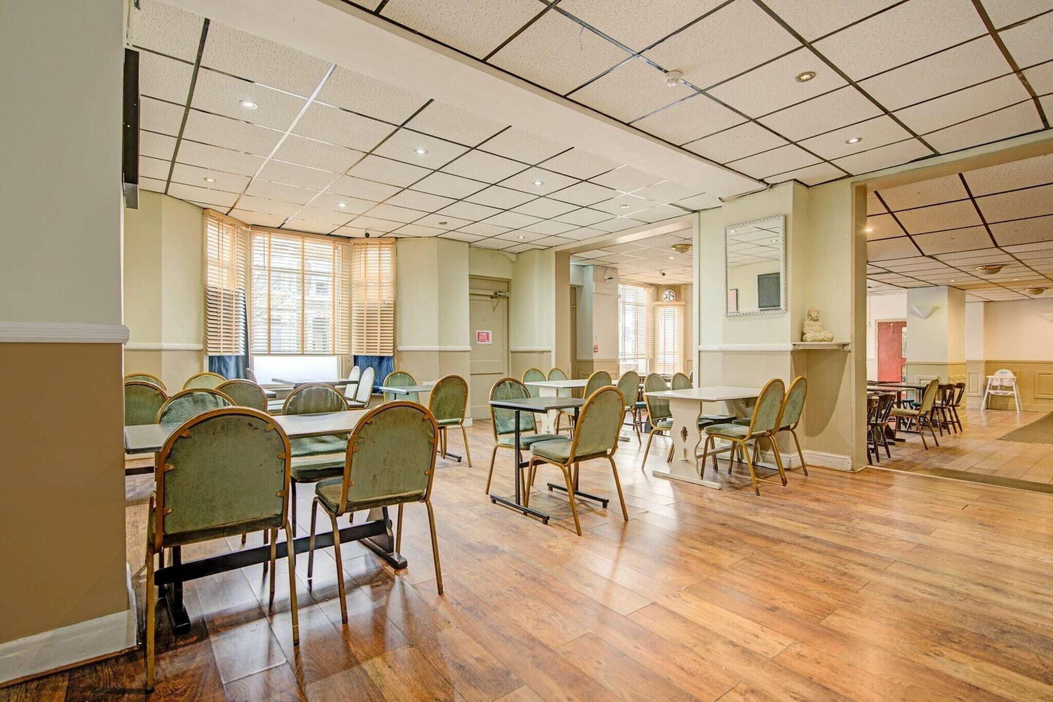 A spacious room with multiple tables and chairs, large windows with blinds, and a tiled ceiling.