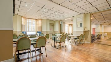 A spacious room with multiple tables and chairs, large windows with blinds, and a tiled ceiling.
