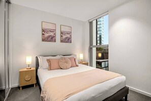 Ein Schlafzimmer mit einem großen Bett, zwei Nachttischlampen, einem Fenster mit Blick auf ein Gebäude und zwei gerahmten Bildern an der Wand.