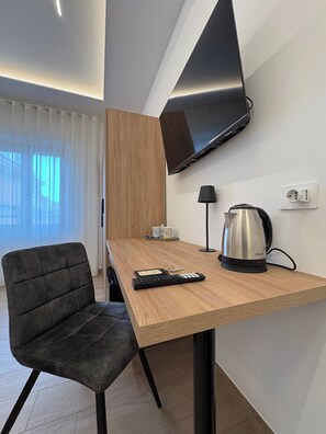 A modern hotel room with a wooden desk, a black chair, a flat-screen TV mounted on the wall, a coffee maker, and a lamp.