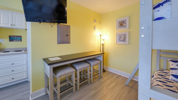 A room with a TV mounted on the wall, a desk with three chairs, a bed with a patterned pillow, and a sign on the wall that says "Good Beach." - Walk to Boardwalk & Beach! Ocean City Studio (Ocean City)