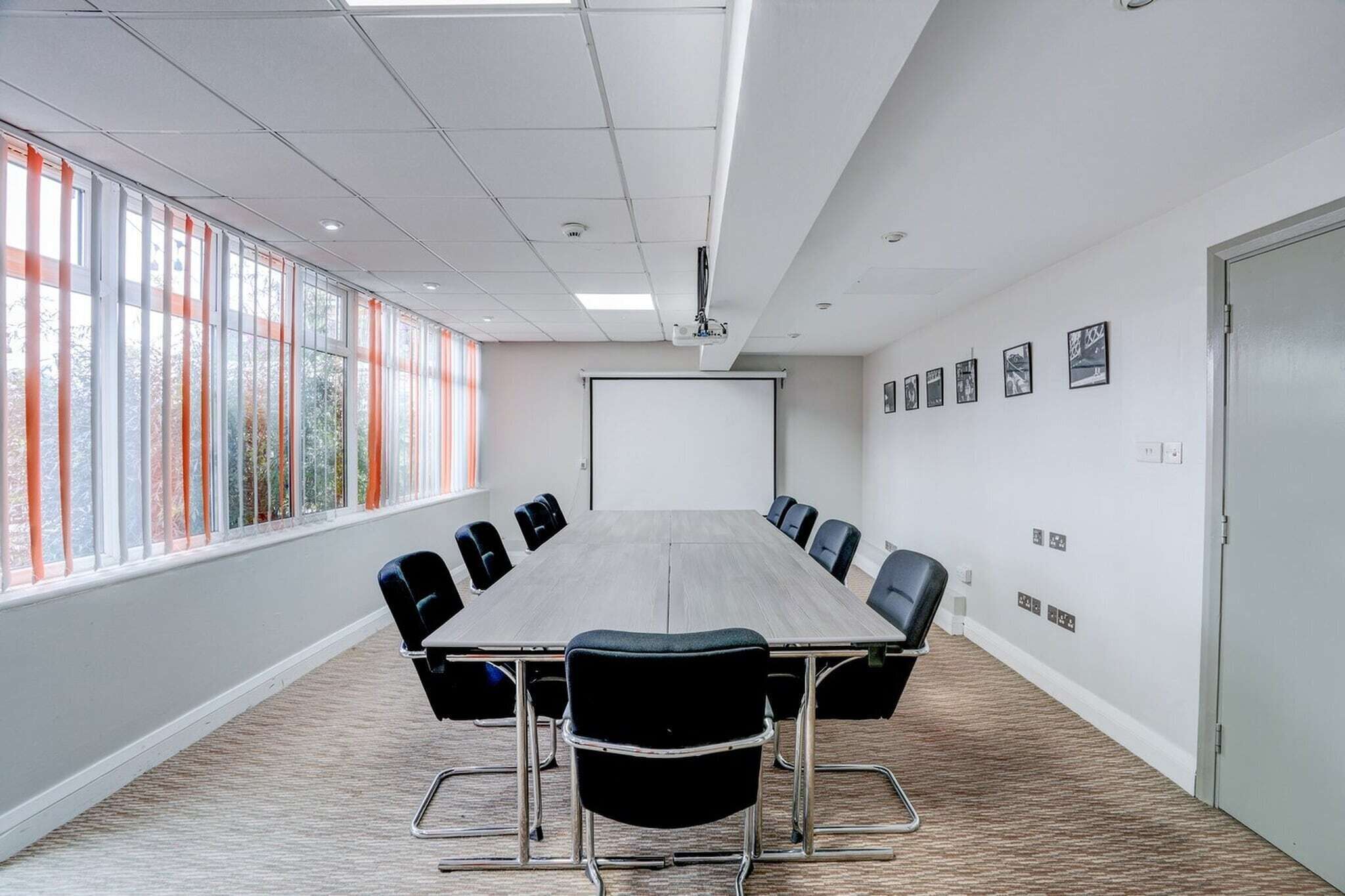 A conference room with a long table, chairs, a projector screen, and large windows with blinds.