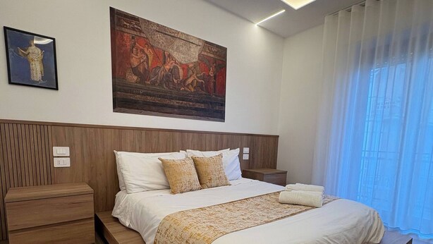 A modern bedroom with a large bed, wooden headboard, a nightstand, a bedside table, and a painting on the wall. - Suite with balcony near Pompeii, Sorrento, and Amalfi (Castellammare di Stabia)