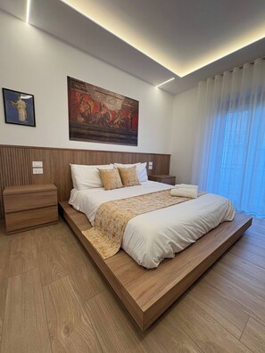 A modern bedroom with a large bed, wooden headboard, a nightstand, a bedside table, and a painting on the wall.