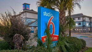 A large blue sign with 'studio 6' and 'stay' text, surrounded by greenery and a building in the background.