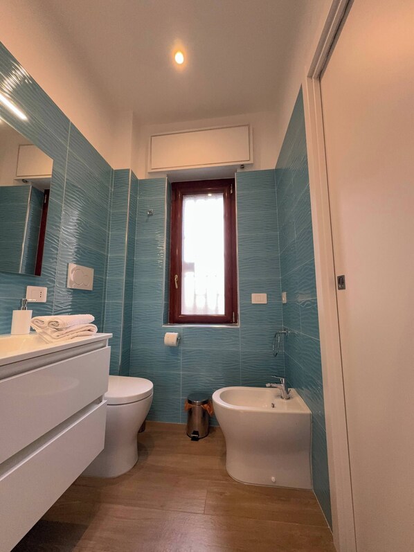 Ein Badezimmer mit Toilette, Bidet und Waschbecken, mit blauen Fliesen verziert, mit Fenster und Spiegel über dem Waschbecken.