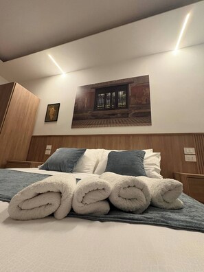Een keurig opgemaakt bed met opgerolde handdoeken, een ingelijst foto aan de muur en een houten garderobe.