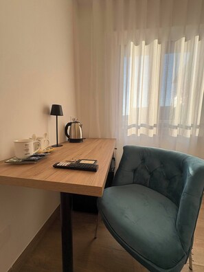 A room with a wooden table, a blue chair, a coffee maker, a lamp, and a window with sheer curtains.