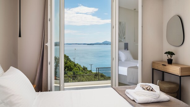 A bedroom with a bed, a bedside table with a plant, a mirror, and a view of the sea through an open door. - Aithra Seaview Suite by Konnect (Barbati)