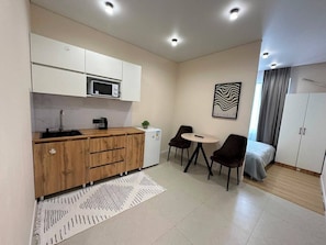 Cocina compacta equipada con microondas, lavamanos y refrigerador pequeño.  Zona de comedor con dos sillas y una mesa. La habitación tiene una cama con cubrecama gris y una ventana con cortinas.