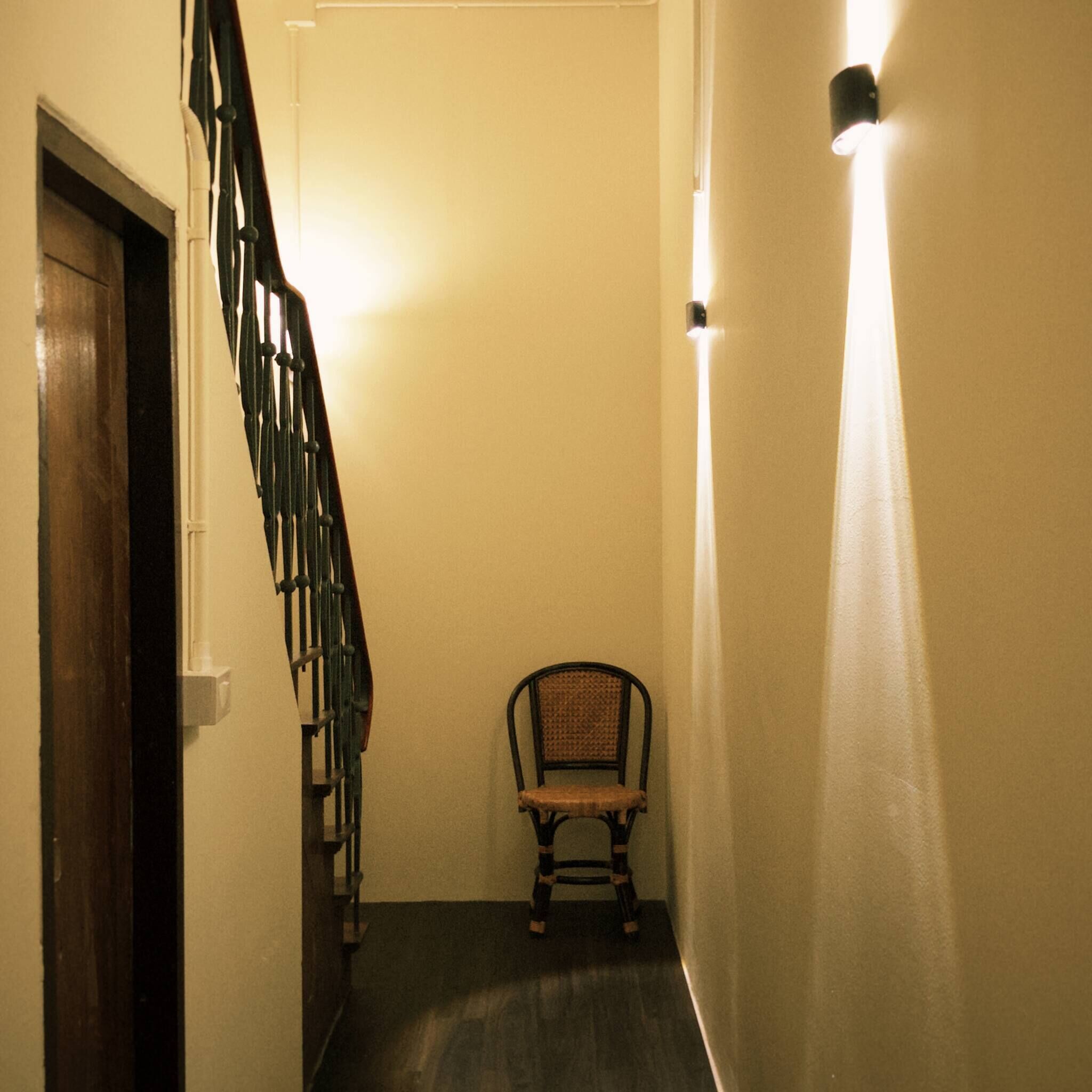 A narrow hallway with a wooden staircase on the left, a chair in the center, and a wall-mounted light on the right.