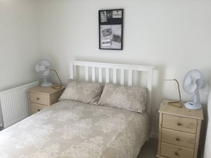 Une chambre à coucher comprenant un lit, deux tables de chevet, deux ventilateurs fixés au mur, un radiateur et un tableau encadré accroché au mur.