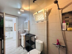 Un baño moderno con inodoro, lavabo, espejo, ducha y un estante con artículos de aseo.