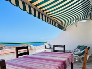 Un balcón con toldo a rayas, una mesa con mantel estampado, una cama con almohadas y vistas al mar.