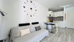 Ein modernes Wohnzimmer mit einer Couch, einer Wanduhr, einer Küchenzeile mit Mikrowelle und einem Esstisch.