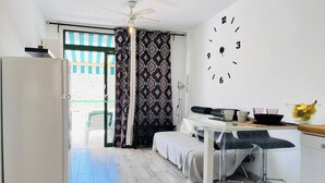 Ein kleines Zimmer mit einem Bett, einem kleinen Tisch mit Stuhl, einem Kühlschrank, einer Wanduhr und einem Fenster mit Vorhängen.