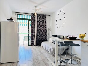 Ein kleines Zimmer mit einem Bett, einem kleinen Tisch mit Stuhl, einem Kühlschrank, einer Wanduhr und einem Fenster mit Vorhängen.