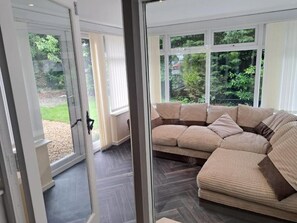 A modern living room with a large sectional sofa, a glass door leading outside, and a view of greenery. - Charming 4-Bedroom Holiday Home in  Chorley (Chorley)