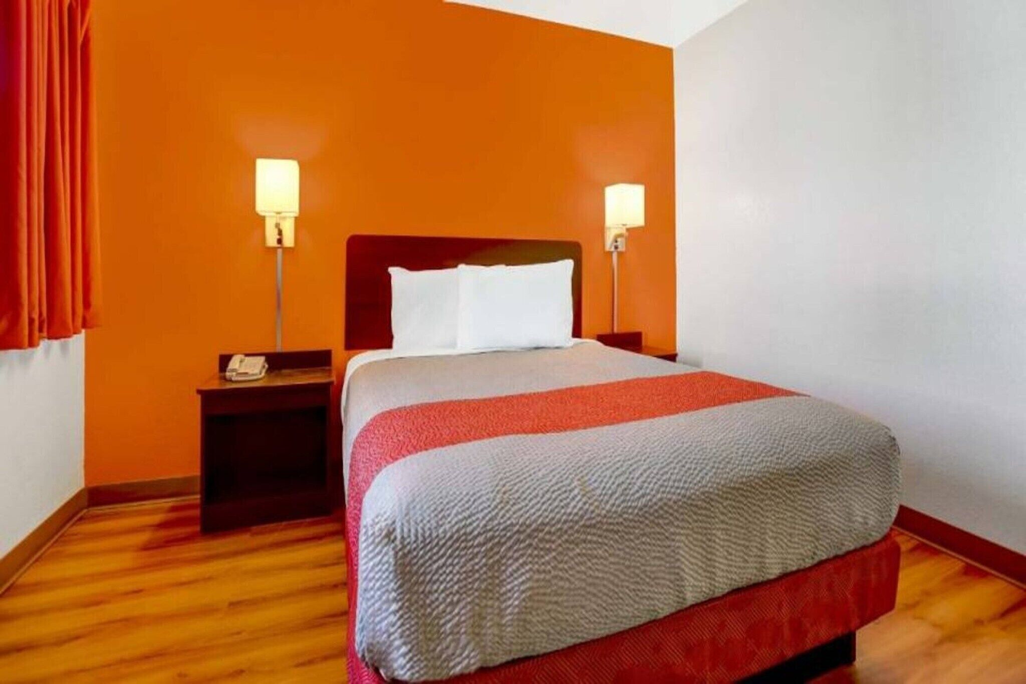 Quarto de hotel com cama, duas luminárias de cabeceira, mesa de apoio e piso de madeira.