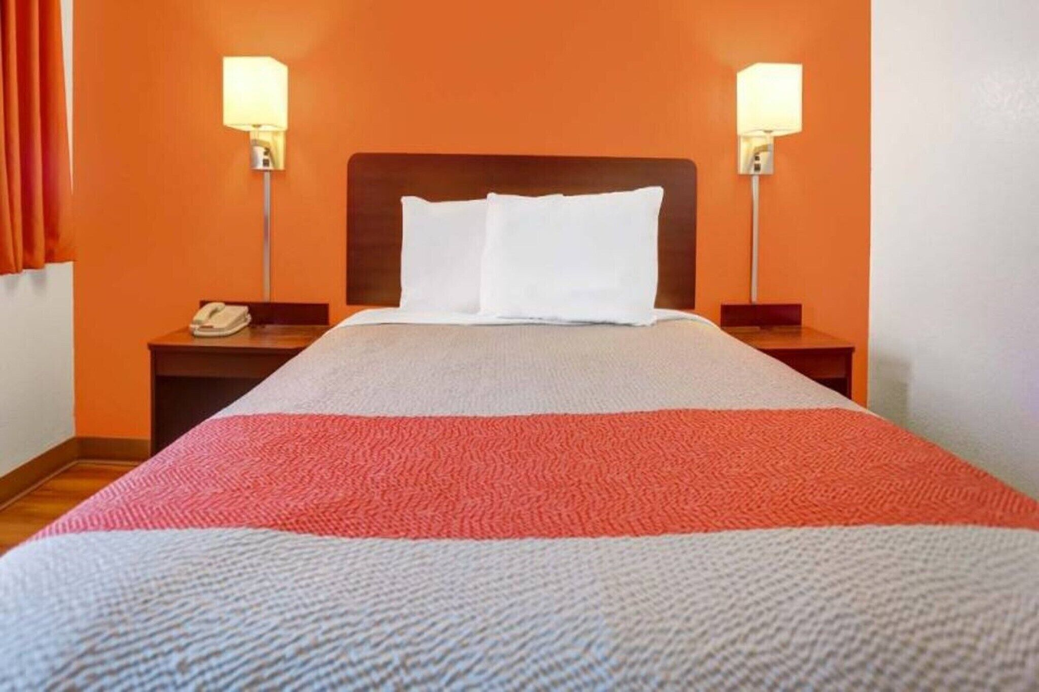 Quarto de hotel com uma cama, duas luminárias de cabeceira, uma mesa de apoio com telefone e uma parede laranja.