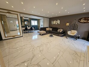 A spacious living area with a marble floor, a dark sofa, a hanging chair, and a dining area with chairs and a table.