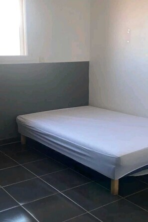 Une petite chambre avec un lit simple, aux couleurs gris et blanc, dotée d’un sol carrelé et d’une fenêtre située sur le côté gauche.