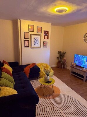 Ein Wohnzimmer mit einem dunkelgrauen Sofa, einem kleinen runden Tisch mit einer Vase und einem Fernseher auf einem Schrank.