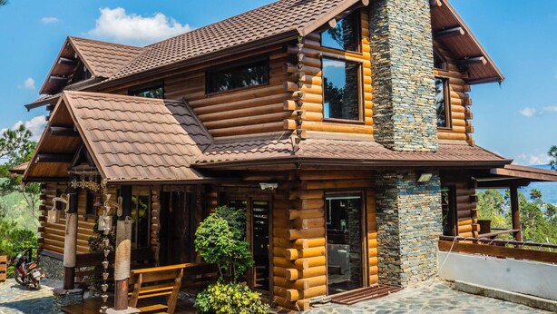 Eine Blockhütte mit einem Steinsschornstein, großen Fenstern und einer Terrasse mit Steinplatten.