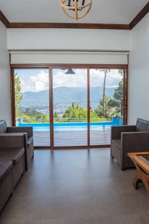 Ein Zimmer mit einer großen Schiebeglaswand, die zu einem Pool führt, mit Korbmöbeln und Bergblick.