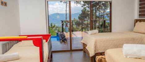 A room with two beds, a red bunk bed, and a large sliding glass door leading to an outdoor deck with a view of mountains.
