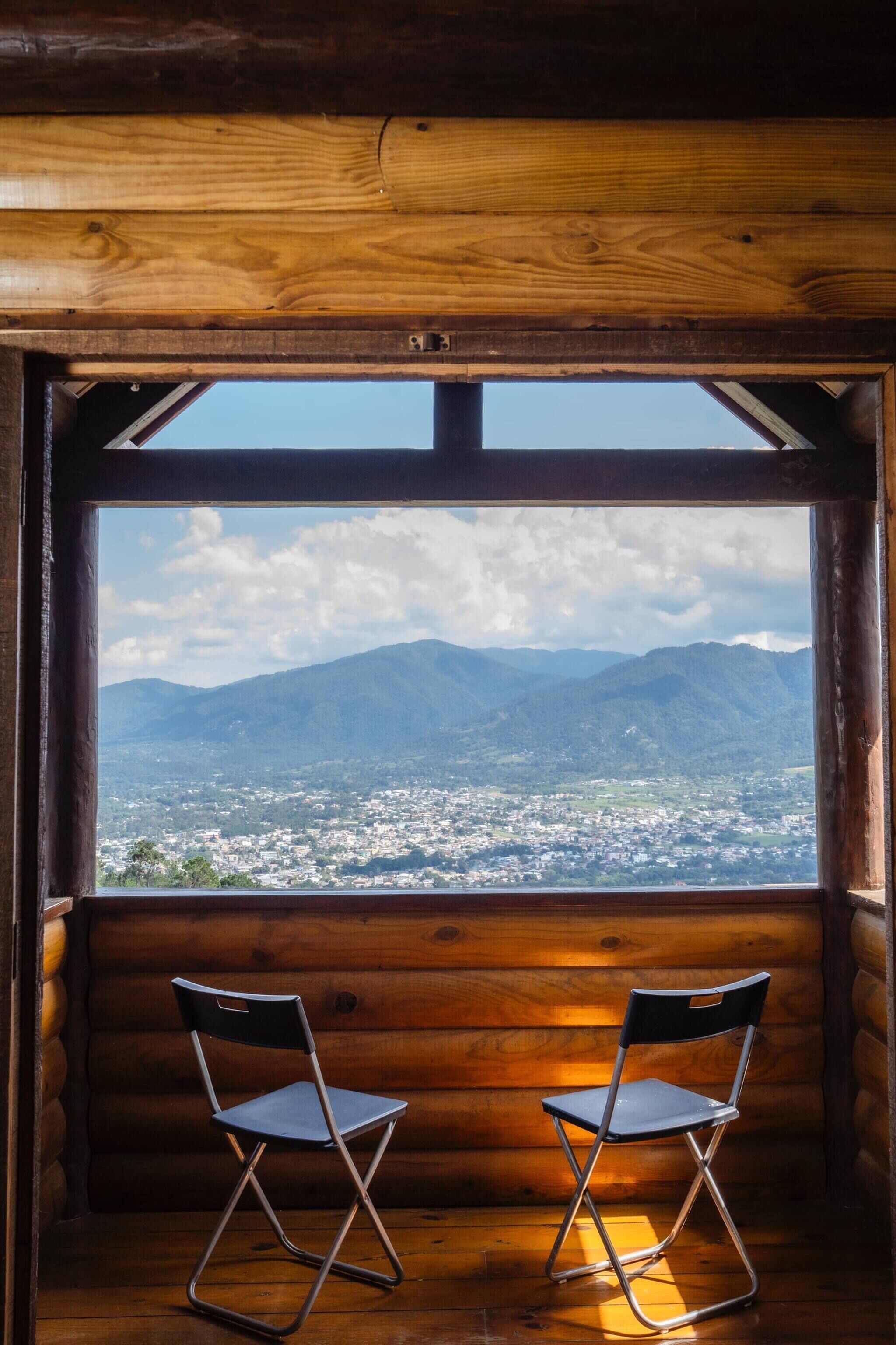 L’intérieur d’une cabane avec un mur en rondins de bois, une fenêtre donnant sur une ville et des montagnes, et deux chaises pliantes.