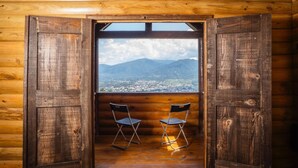 Eine Holzhütte mit einem großen Fenster, zwei Klappstühlen und Blick auf eine Berglandschaft.