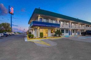 Un motel con un letrero de 'WELCOME', un estacionamiento y un cartel con el número '6' que indica una ubicación cercana.