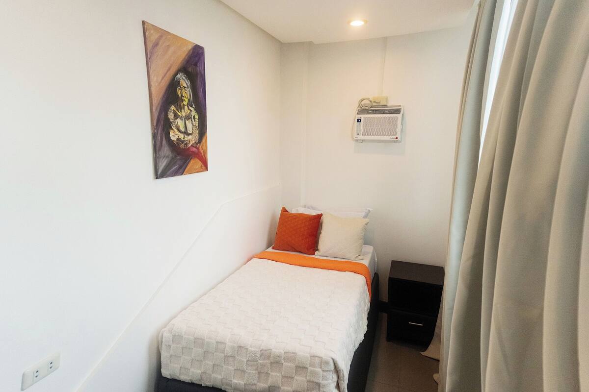 A small, neatly arranged bedroom with a bed, a wall-mounted air conditioner, and a painting.