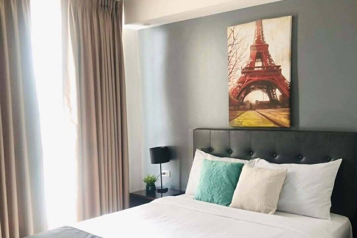 A hotel room with a large bed, a nightstand with a lamp, a painting of the Eiffel Tower, and a window with curtains.