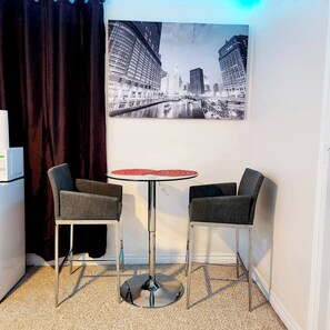 Una habitación con una mesa redonda, dos sillas, un cuadro grande de un paisaje urbano, una cortina oscura y una pared blanca.