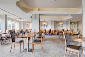 Ampla sala de jantar de hotel, com várias mesas e cadeiras, janelas grandes e luminárias modernas.
