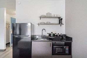 A compact kitchen with a stainless steel refrigerator, a microwave, a sink, and a shelf with dishes.
