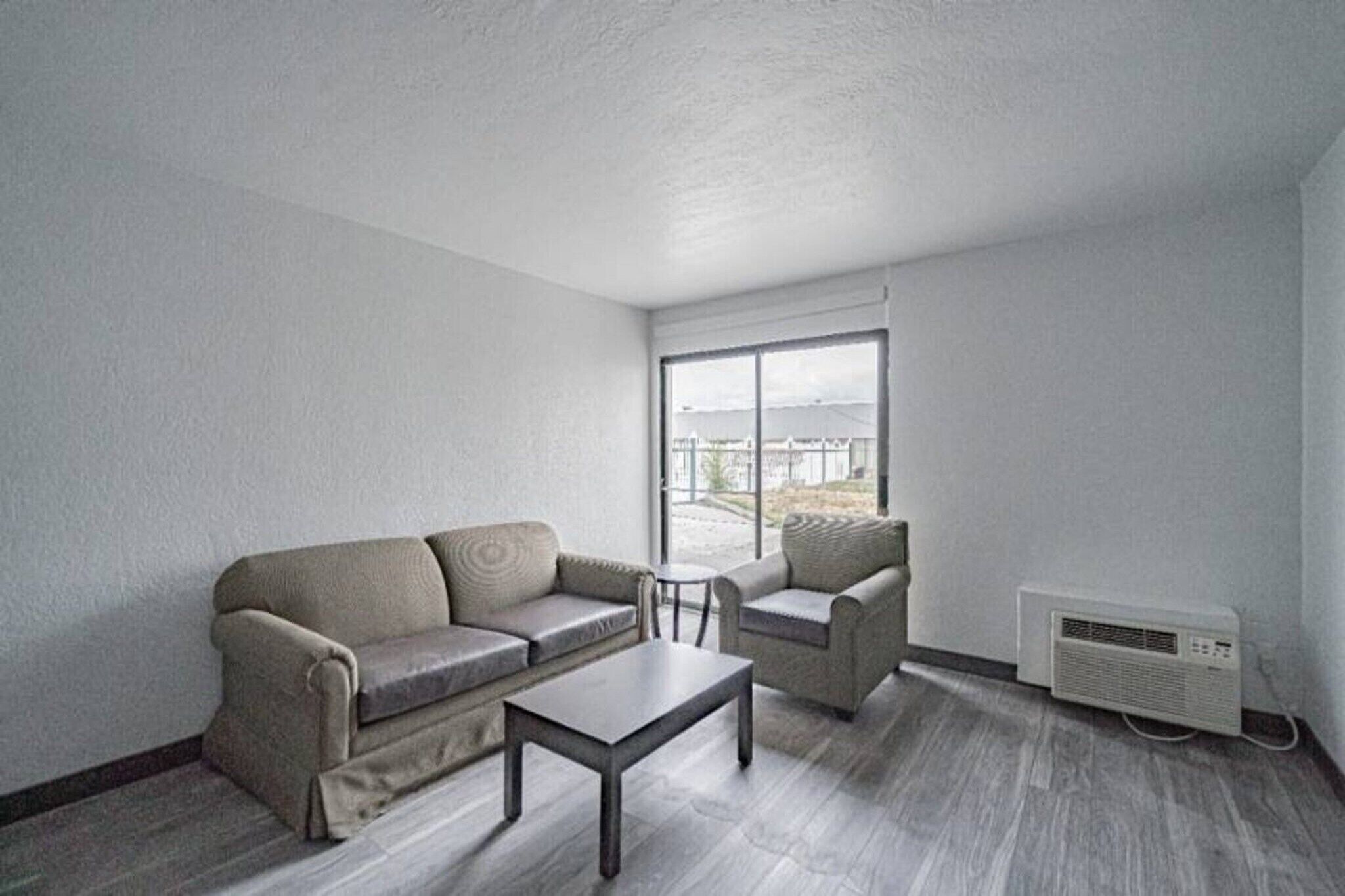 A modern living room with a sofa, armchair, and coffee table, a large window with a view, and a wall-mounted air conditioner.